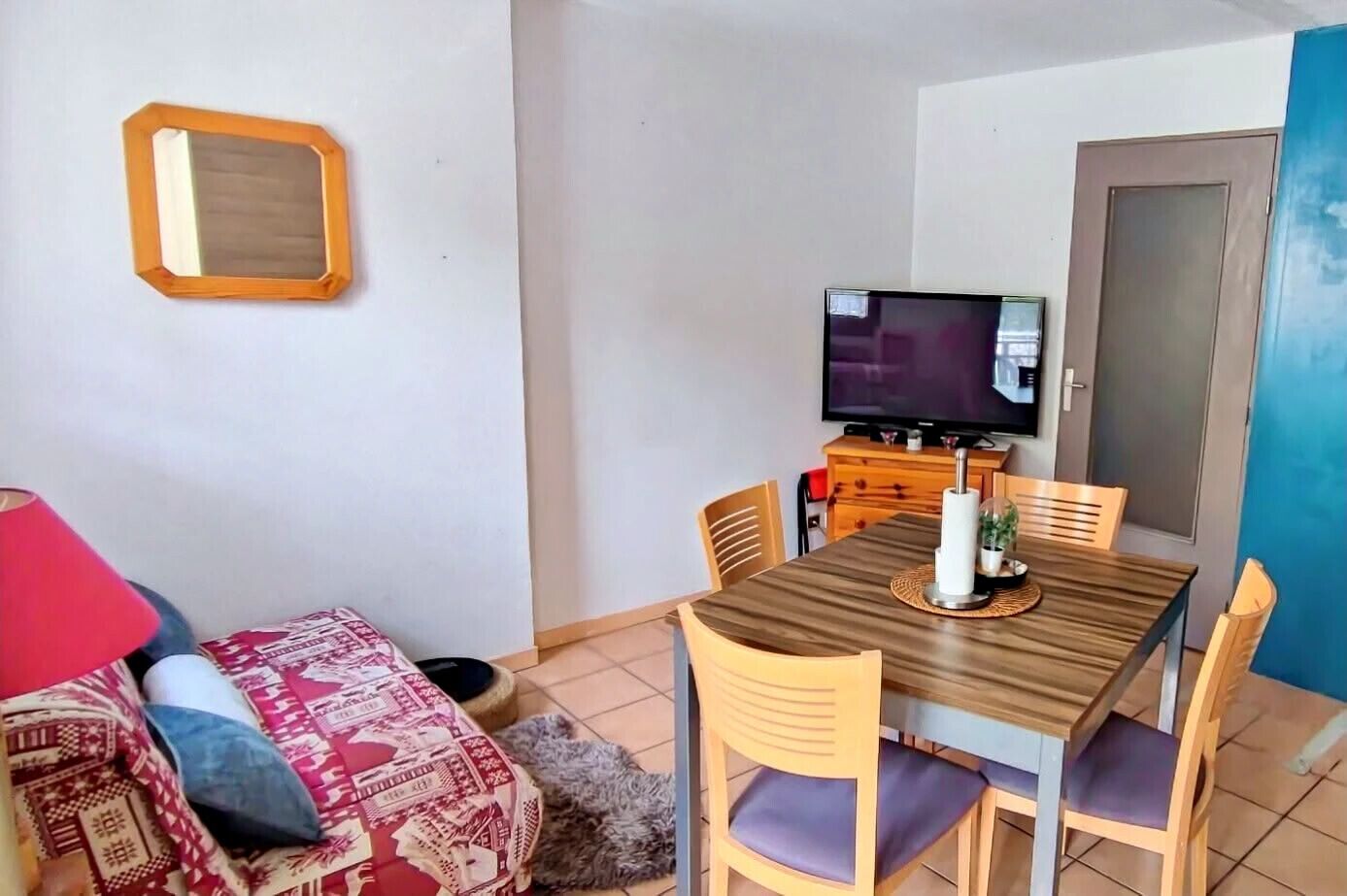 Un pequeño comedor con una mesa de madera, cuatro sillas, un televisor y una lámpara roja.