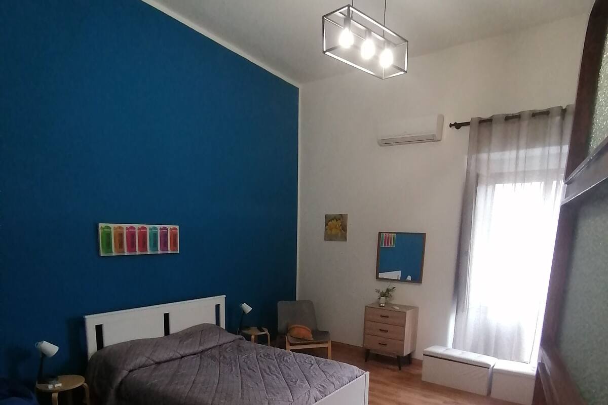 Ein Schlafzimmer mit einem Bett, Nachttischen, einem Schreibtisch, einem Sessel, einem Fenster mit Vorhängen und einer Deckenleuchte.