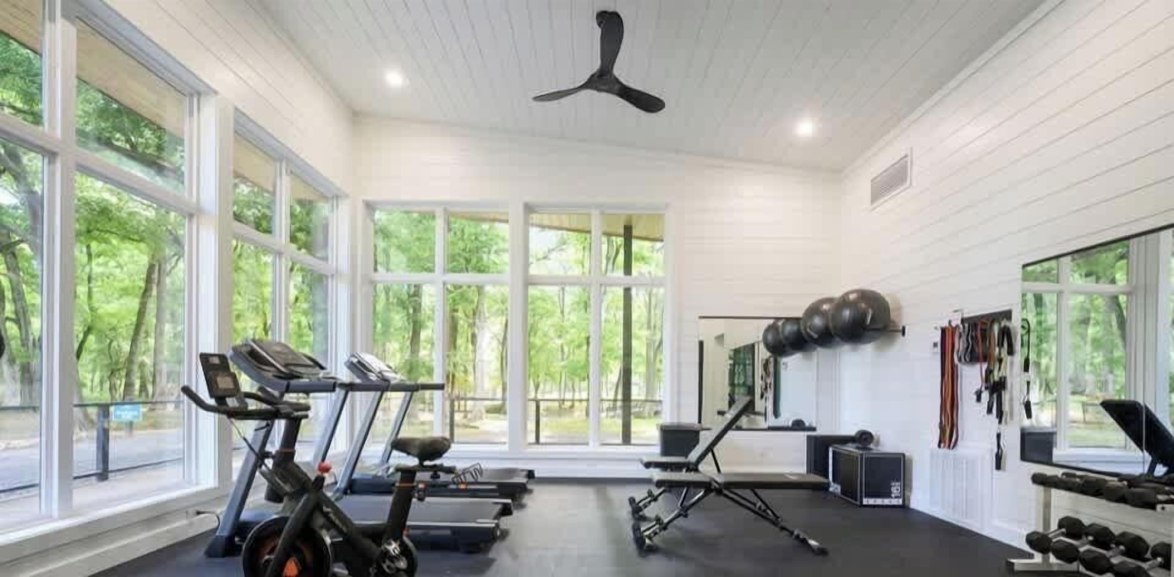 Een sportschool met trainingsapparatuur, grote ramen en een plafondventilator.
