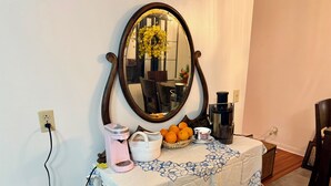 Una pequeña mesa con una cafetera, una cesta de naranjas, una taza y un espejo decorativo en un mueble fijado a la pared.