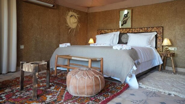A bedroom with a large bed, a wooden bench, a round pouf, and a patterned rug. - Queen Room with Spa Bath # (Agafay)