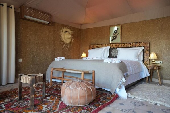A bedroom with a large bed, a wooden bench, a round pouf, and a patterned rug.