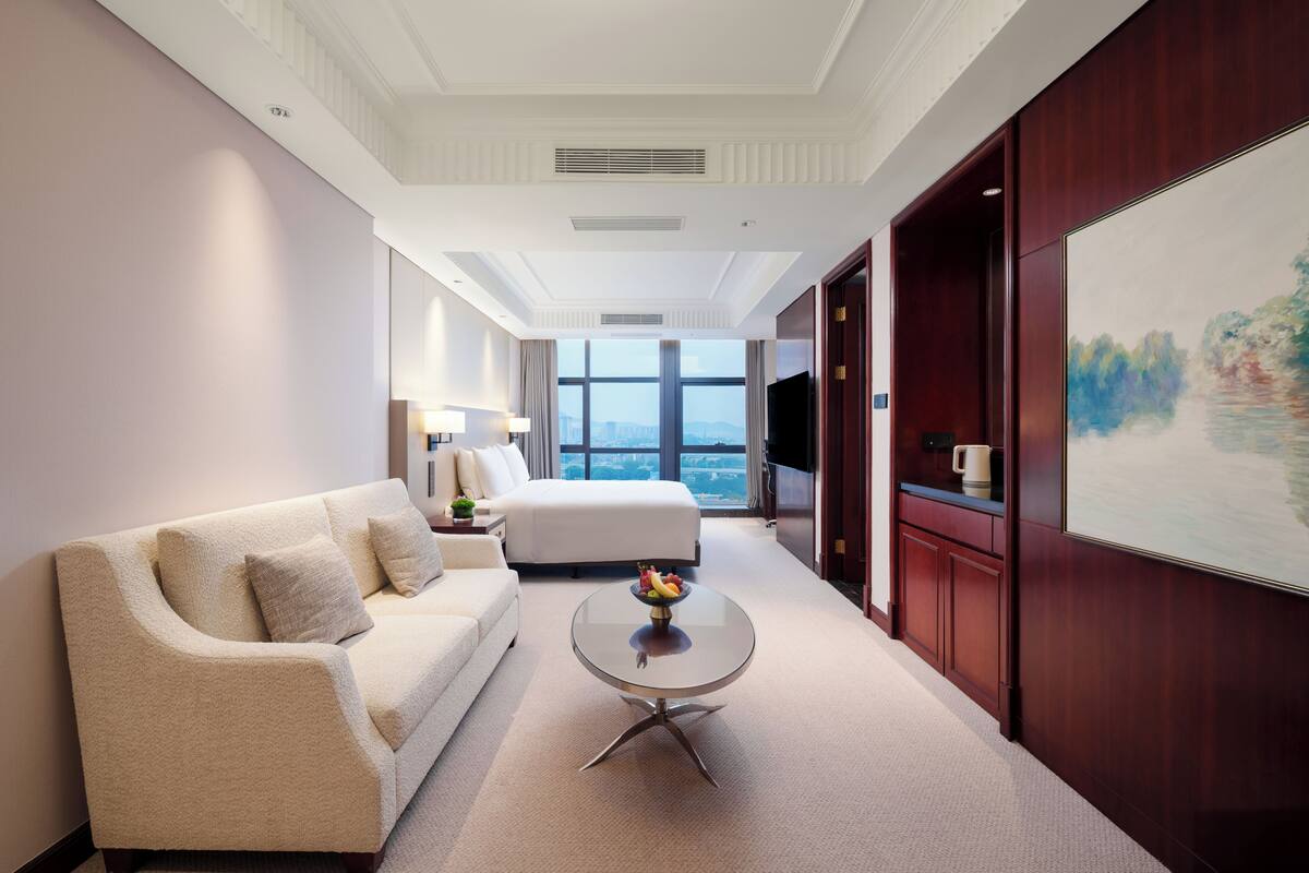Una habitación de hotel con una cama grande, un sofá, una mesa de centro con fruta, un televisor y un cuadro en la pared.