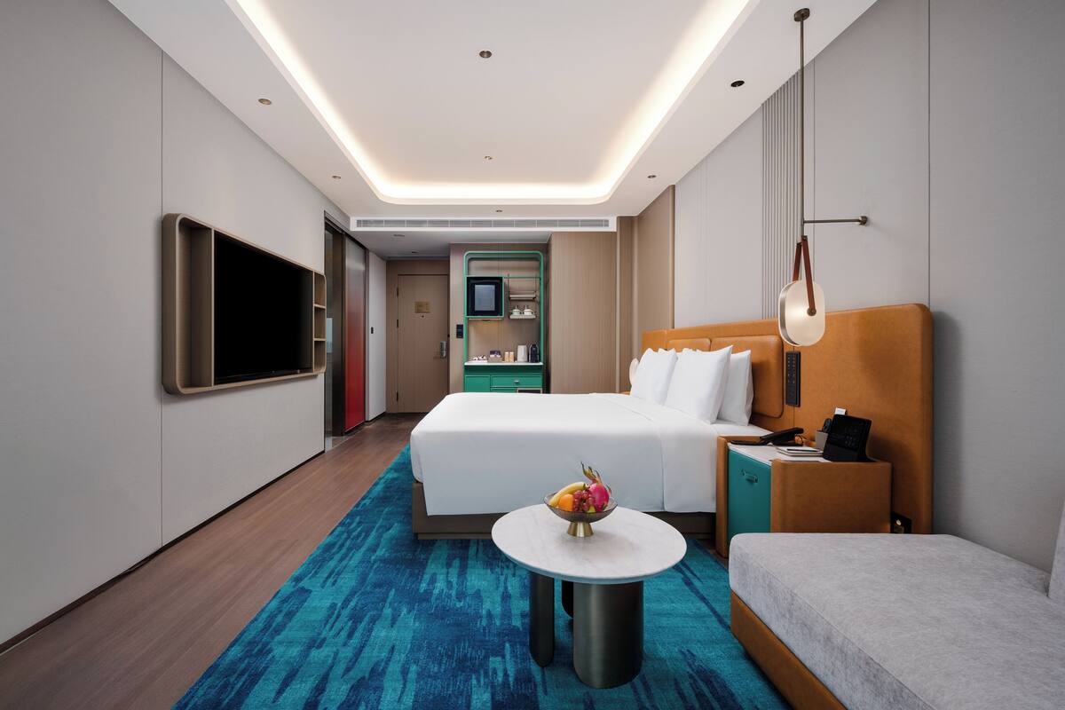 Una habitación de hotel moderna con cama, mesita de noche, lámpara, televisor y una pequeña mesa redonda con fruta.