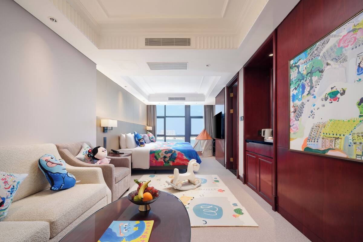 Habitación de hotel con cama, sofá, mesa de centro con frutero, televisor y ventana grande.