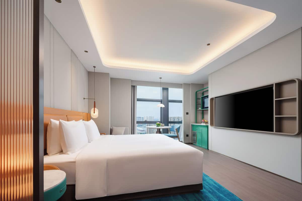 Habitación de hotel moderna con una cama grande, zona de comedor, televisor y vistas a la ciudad.