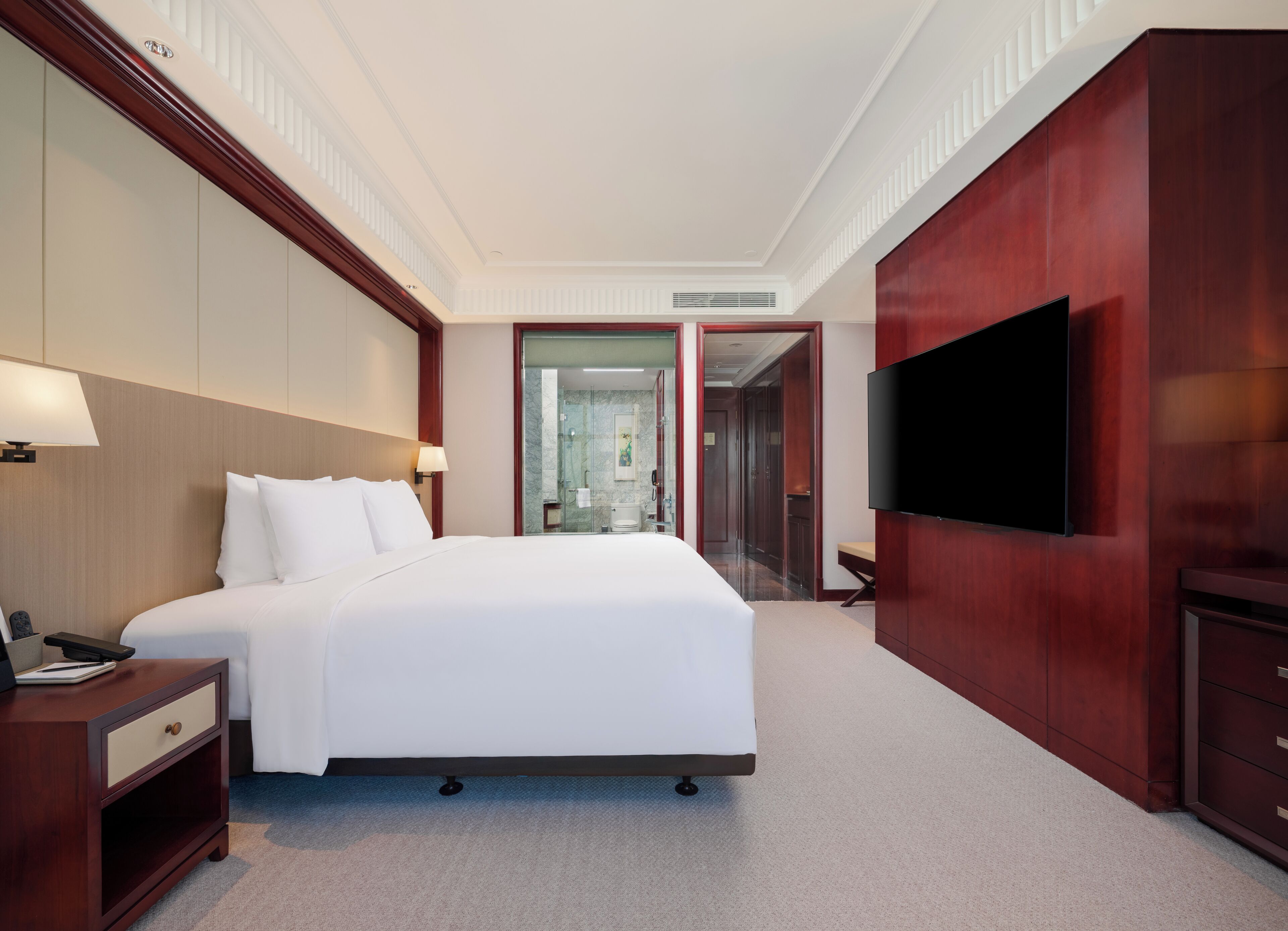 A hotel room with a large bed, bedside tables, a TV, and a bathroom visible through a glass door.