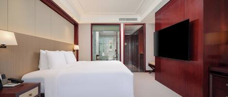 A hotel room with a large bed, bedside tables, a TV, and a bathroom visible through a glass door.