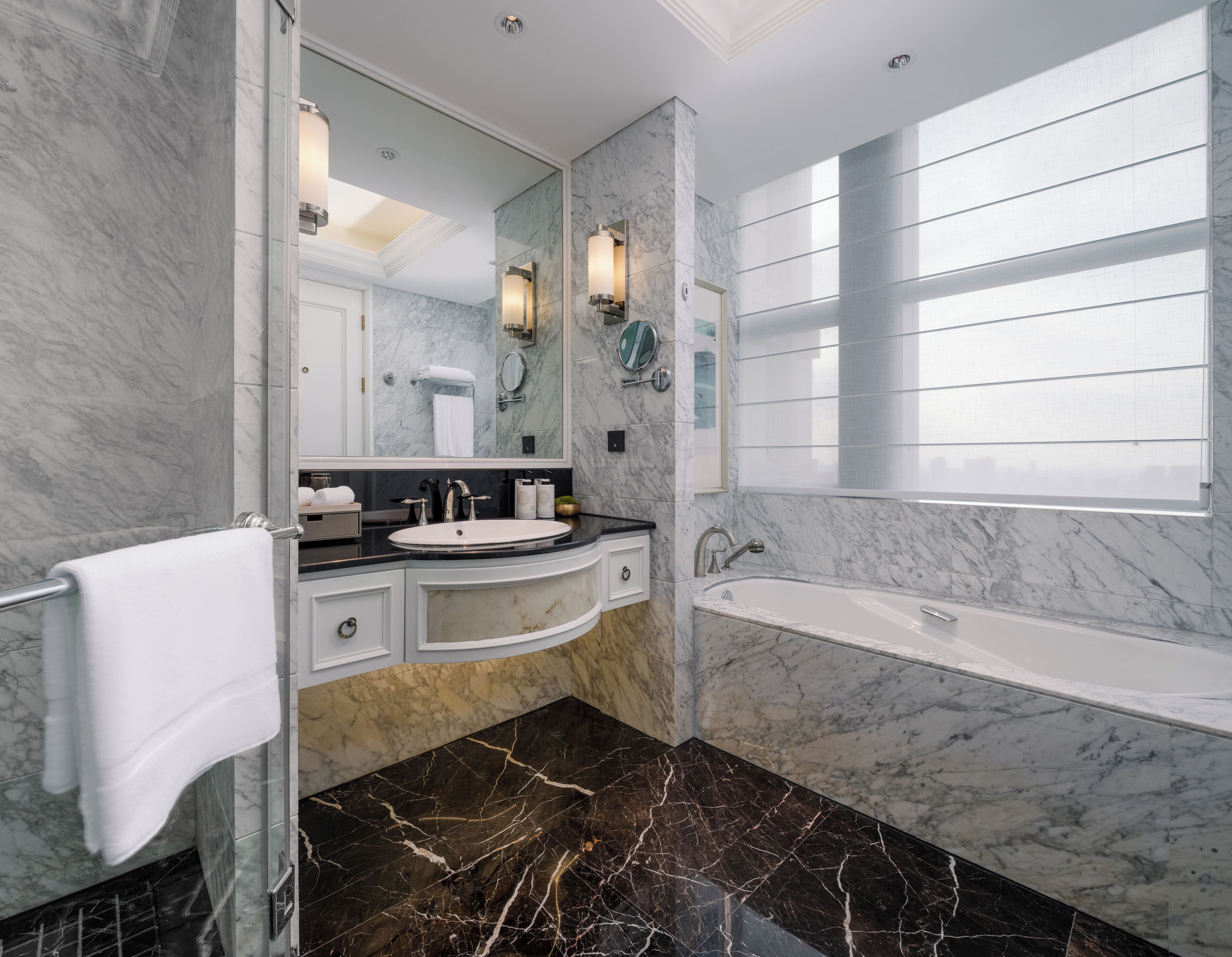 A modern bathroom with a marble vanity, a large mirror, a bathtub, and a window providing natural light.