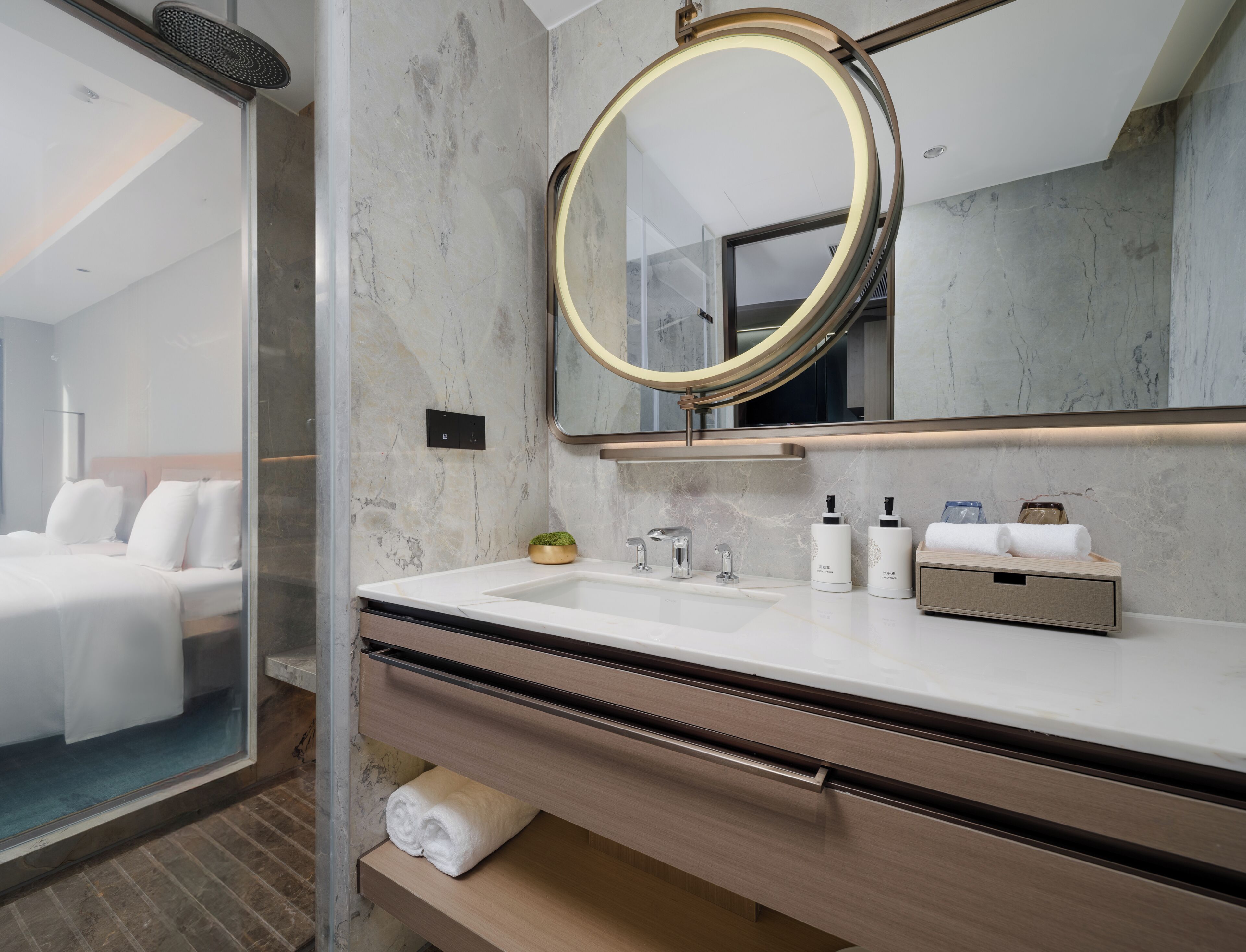 Un baño moderno con pared de mármol, espejo redondo, lavamanos blanco y tocador de madera.