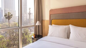 Une chambre d’hôtel avec une grande fenêtre, un lit avec une literie blanche, une table de chevet avec une lampe et une vue sur le paysage urbain.