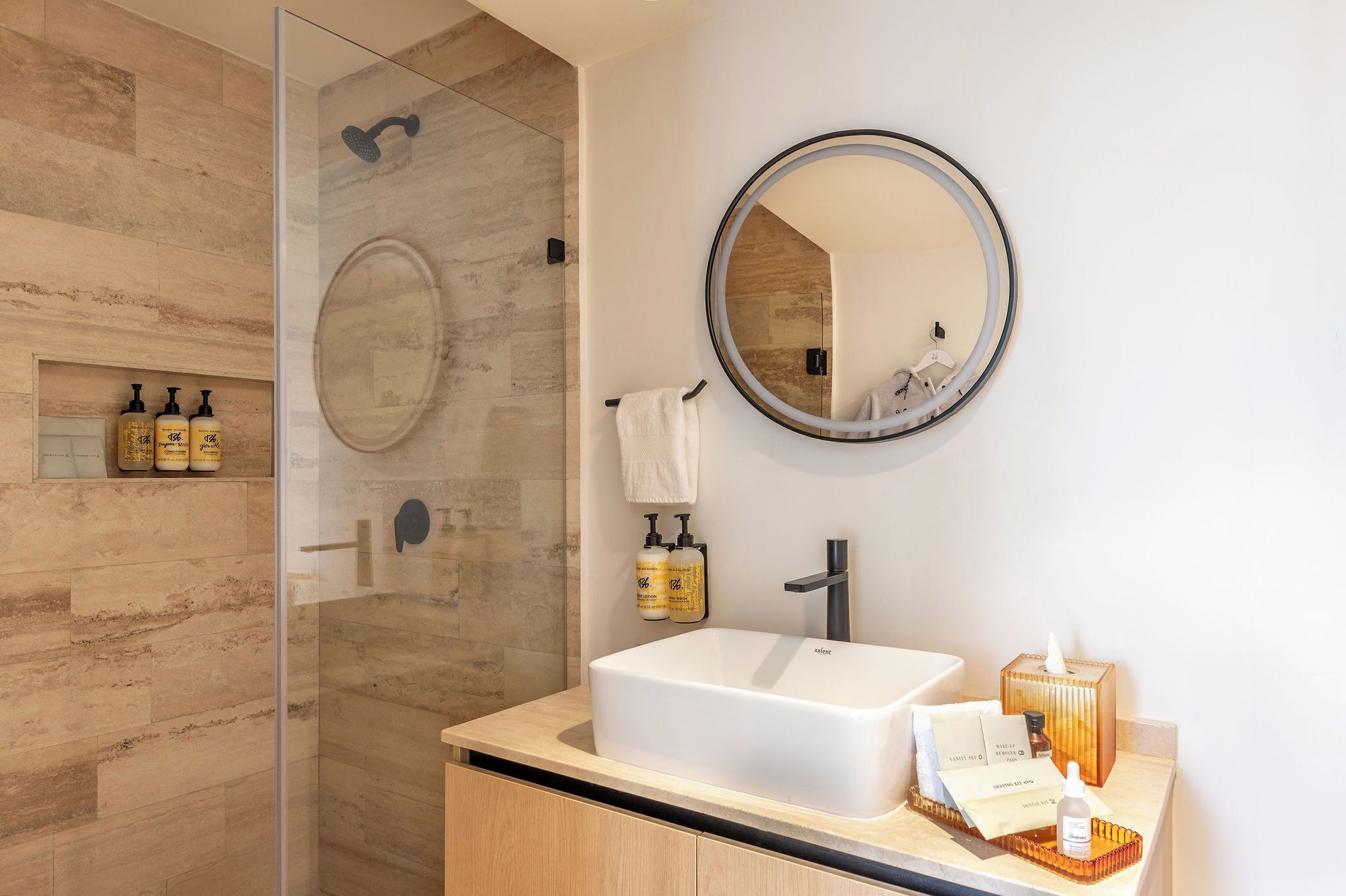 Une salle de bain moderne avec une douche à parois de verre, un grand lavabo, un miroir rond et des articles de toilette disposés sur le comptoir.