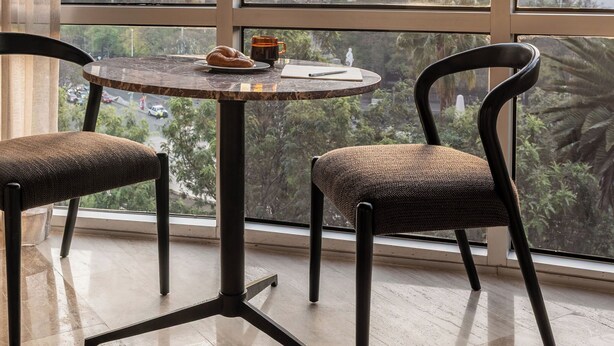 Une pièce avec une grande fenêtre, deux chaises, une petite table ronde avec une tasse et une pâtisserie, et une vue sur les bâtiments de la ville.