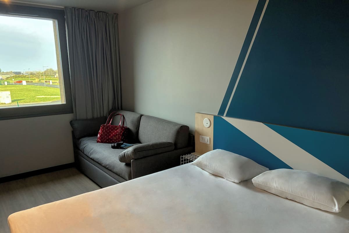 Une chambre d’hôtel avec un lit, un canapé, un sac à main rouge et une vue sur l’extérieur.