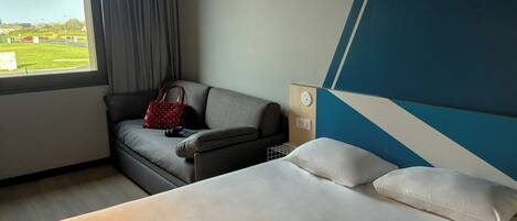 A hotel room with a bed, a sofa, a red handbag, and a view of the outdoors.
