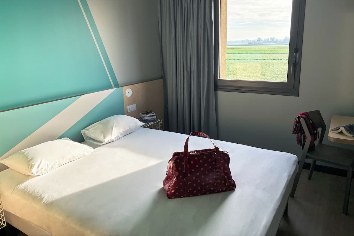 Une chambre d’hôtel avec un lit, une fenêtre donnant sur un champ verdoyant, un bureau avec une chaise et un sac rouge à pois posé sur le lit.