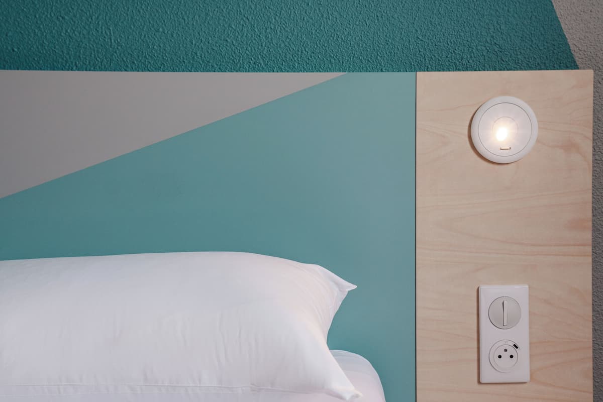 Un lit avec une literie blanche, un mur aux tons bleu sarcelle et gris, et une tête de lit en bois avec un interrupteur et une prise électrique.