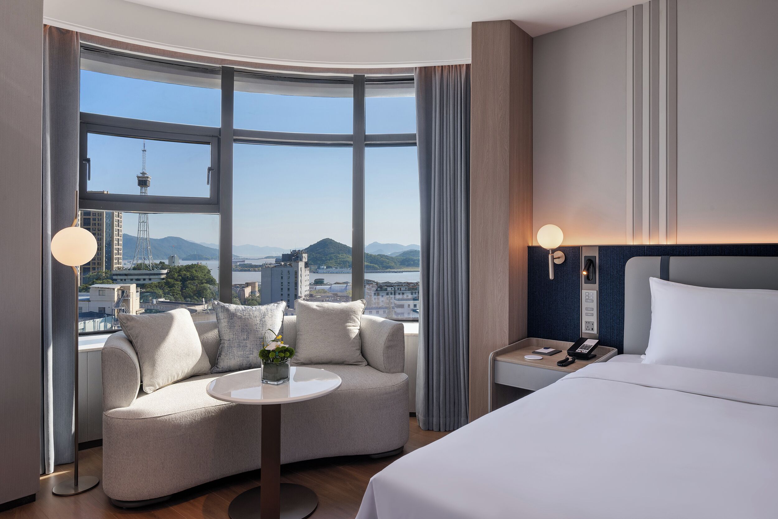 A modern hotel room with a curved sofa, a round table, a bed, and a view of a cityscape through large windows.