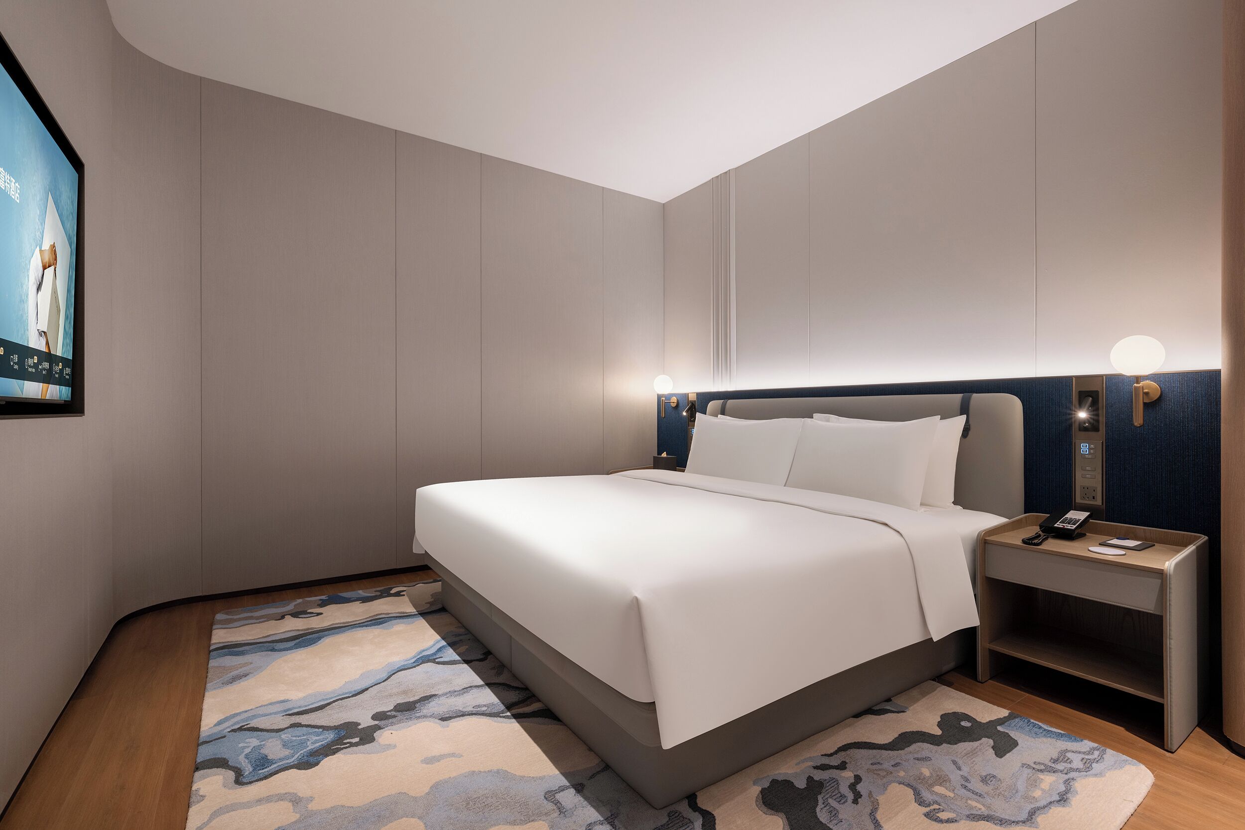 A modern hotel room with a large bed, a bedside table with a lamp, and a wall-mounted TV.