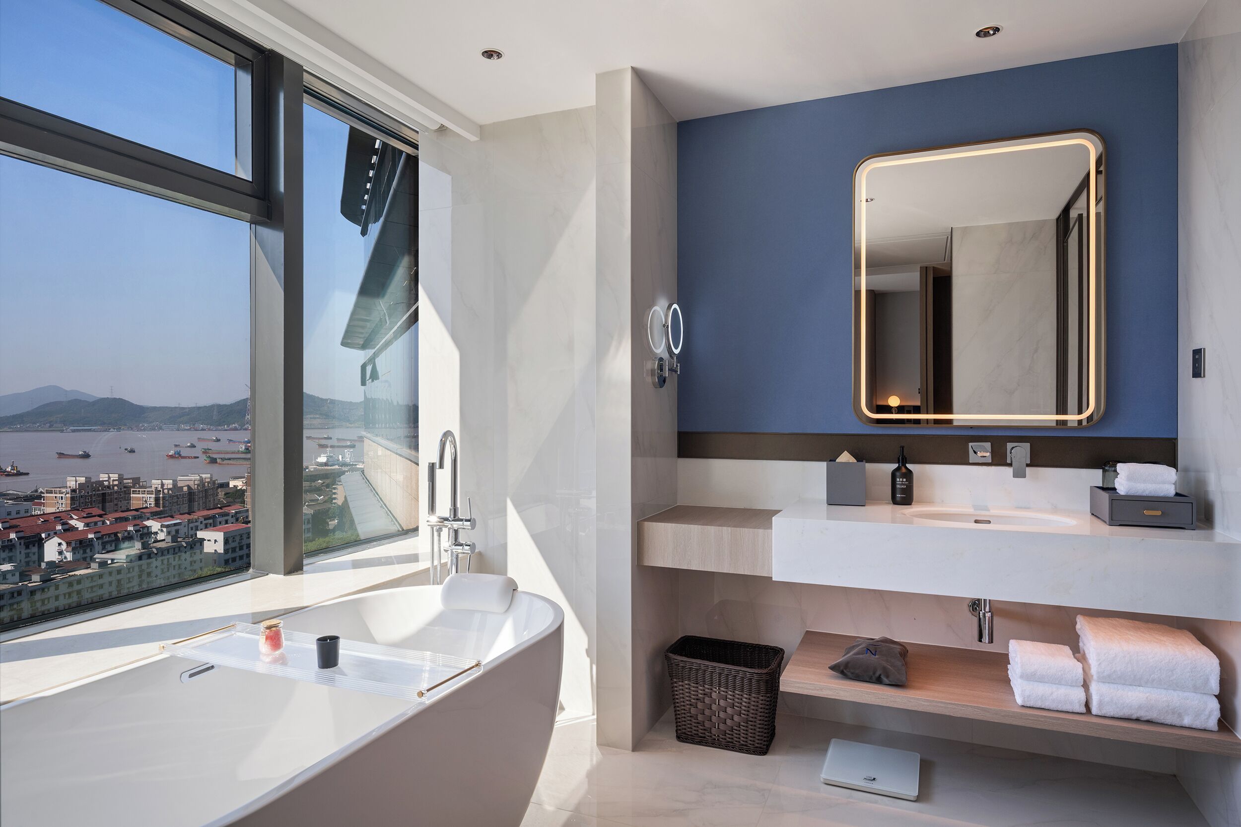 A modern bathroom with a large bathtub, a double sink, and a large mirror. There is a view of a cityscape through a large window.