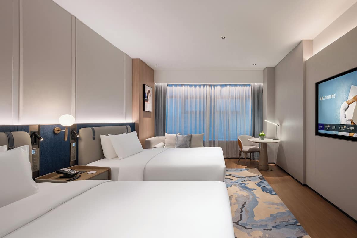 A hotel room with two beds, a desk, a TV, and a large window with curtains.