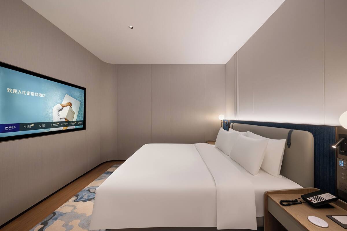 A modern hotel room with a large bed, a flat-screen TV mounted on the wall, a bedside table with a lamp, and a wooden nightstand.