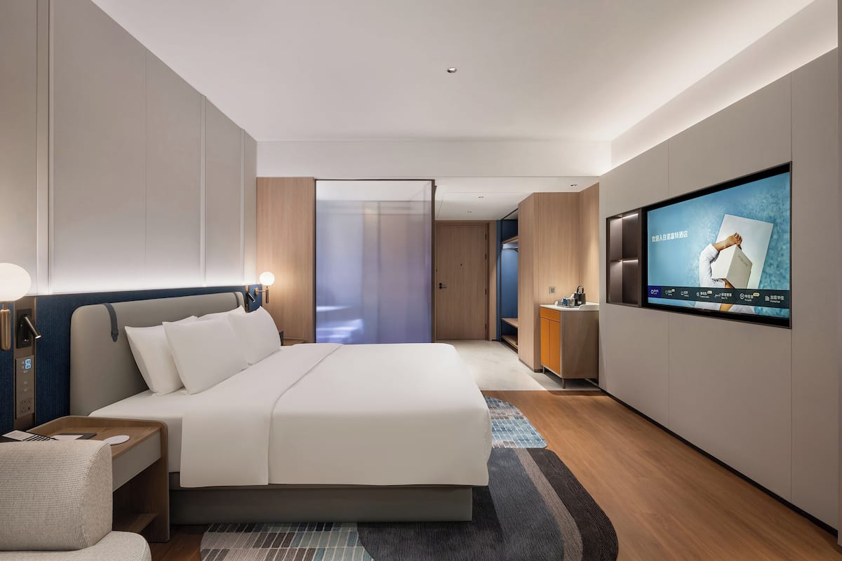 A modern hotel room with a large bed, a TV mounted on the wall, a bedside table with a lamp, and a view of the hallway.