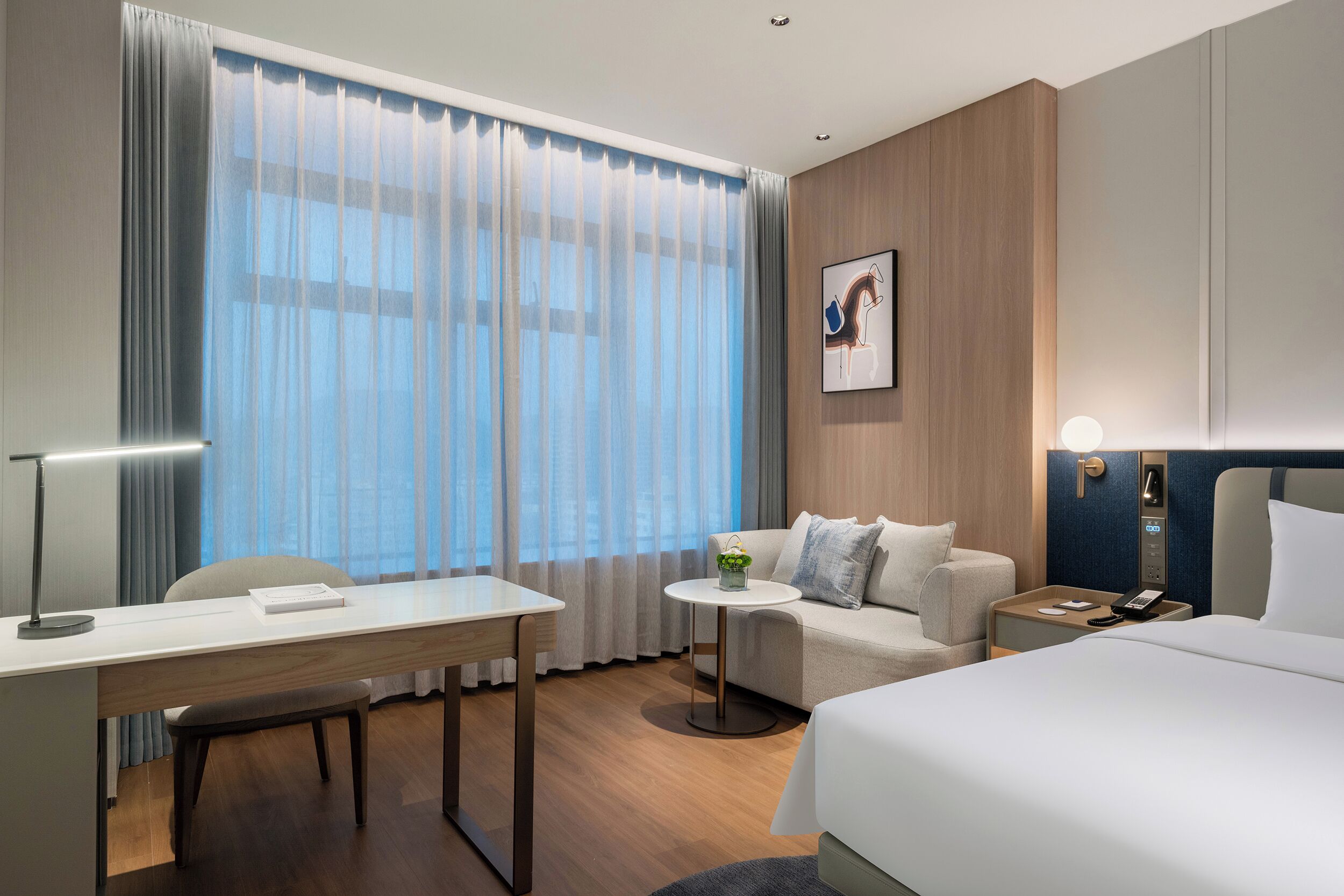 A modern hotel room with a desk, a sofa, a bed, and a small table with a plant.