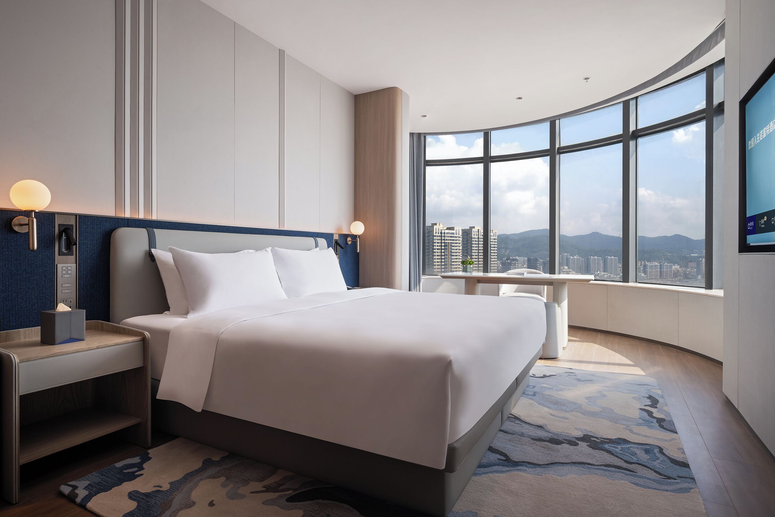 A modern hotel room with a large bed, bedside tables, a desk, and a large window offering a city view.