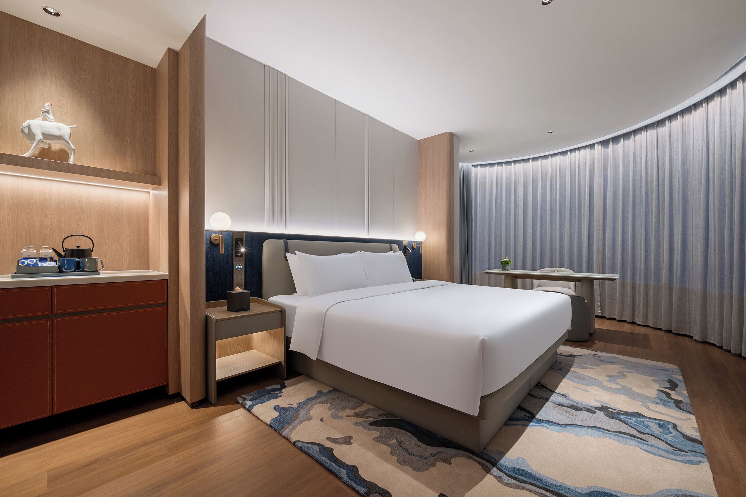 A modern hotel room with a large bed, bedside tables, a desk, and a decorative wall piece.