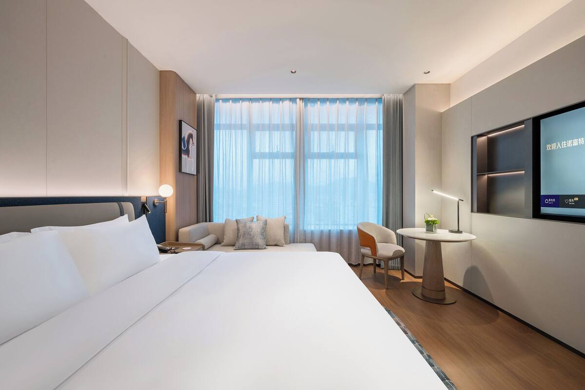 A modern hotel room with a large bed, a desk with a chair, a TV, and a window with curtains.