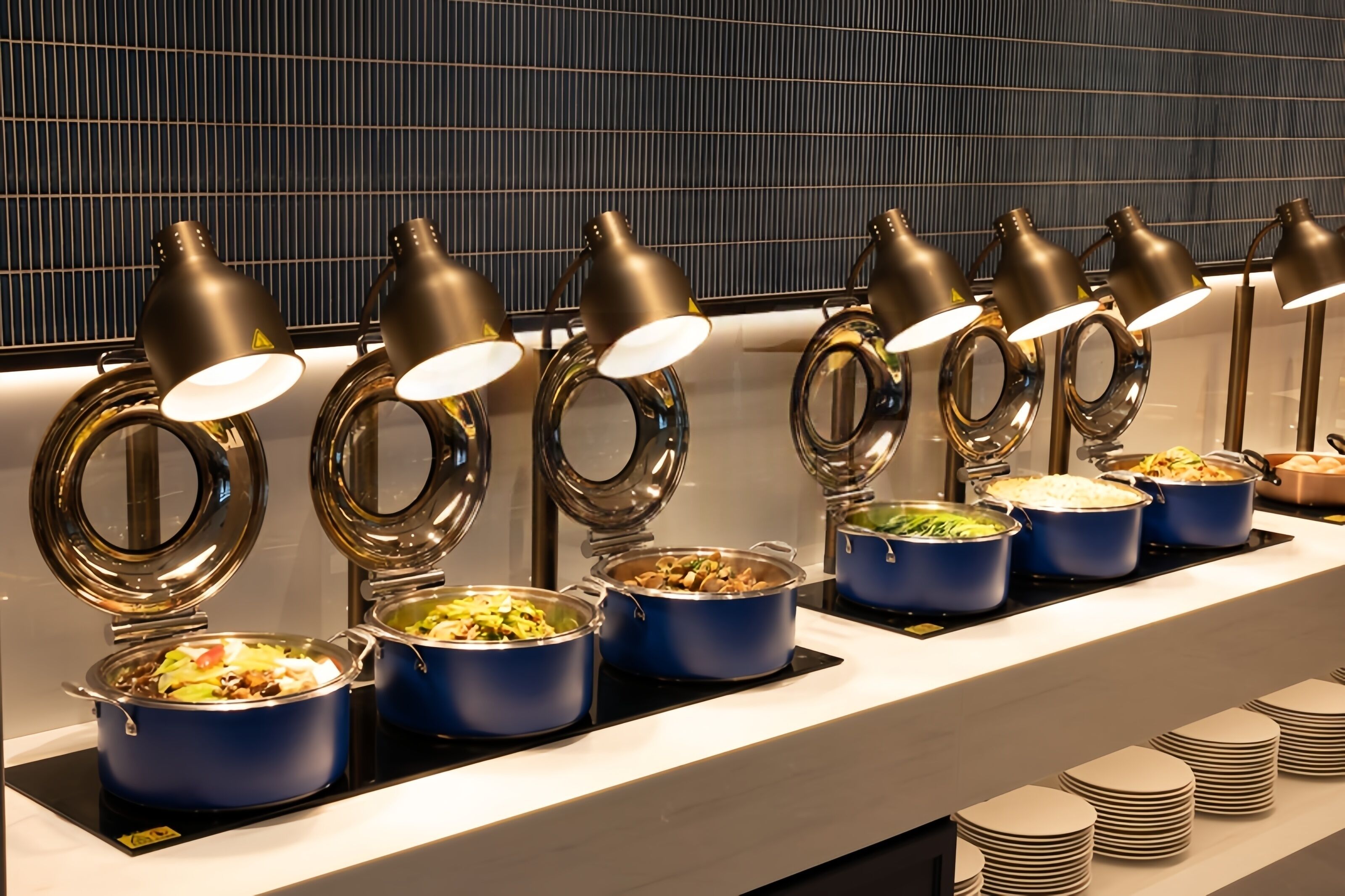 A buffet setup with multiple blue pots of food, black lamps, and stacked plates.