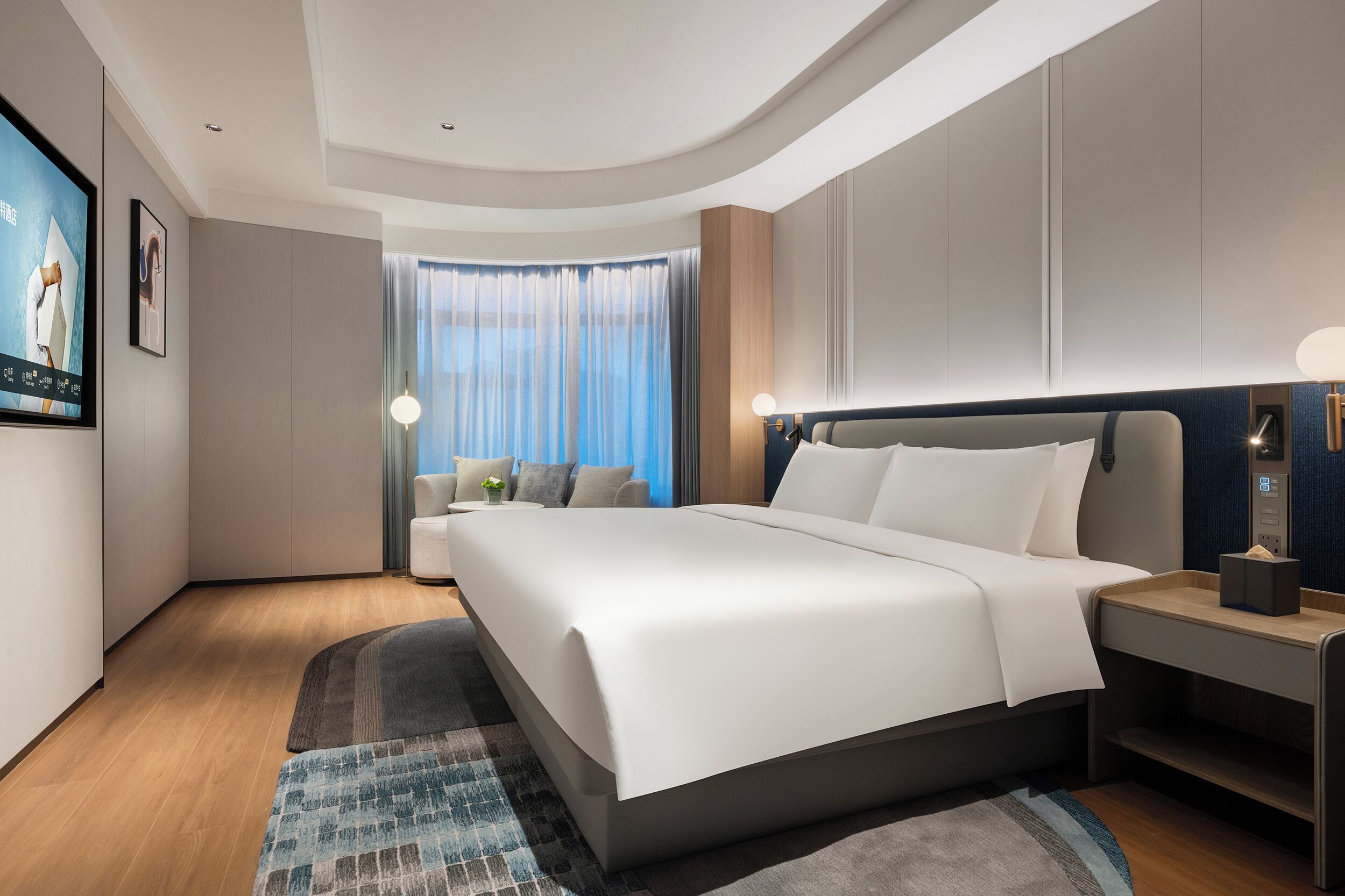 A modern hotel room with a large bed, a bedside table, a nightstand, a TV, and a view of the city through the window.