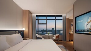 A hotel room with a large bed, a desk, a chair, a TV, and a view of the cityscape through large windows. - Novotel Zhoushan Luting (Zhoushan)