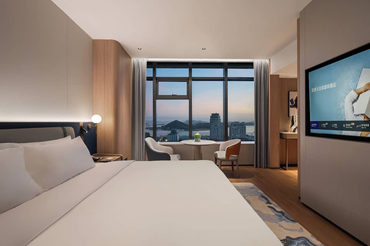 A hotel room with a large bed, a desk, a chair, a TV, and a view of the cityscape through large windows.