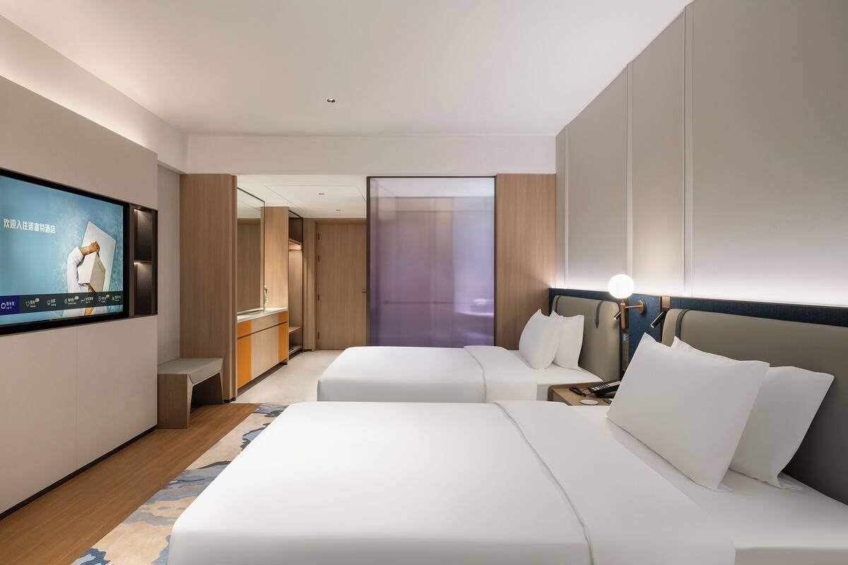A modern hotel room with two beds, a TV, a desk, and a bathroom visible through a door.