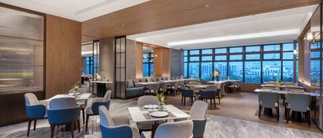A modern dining area with multiple tables and chairs, large windows offering city views, and a high ceiling with wooden paneling.
