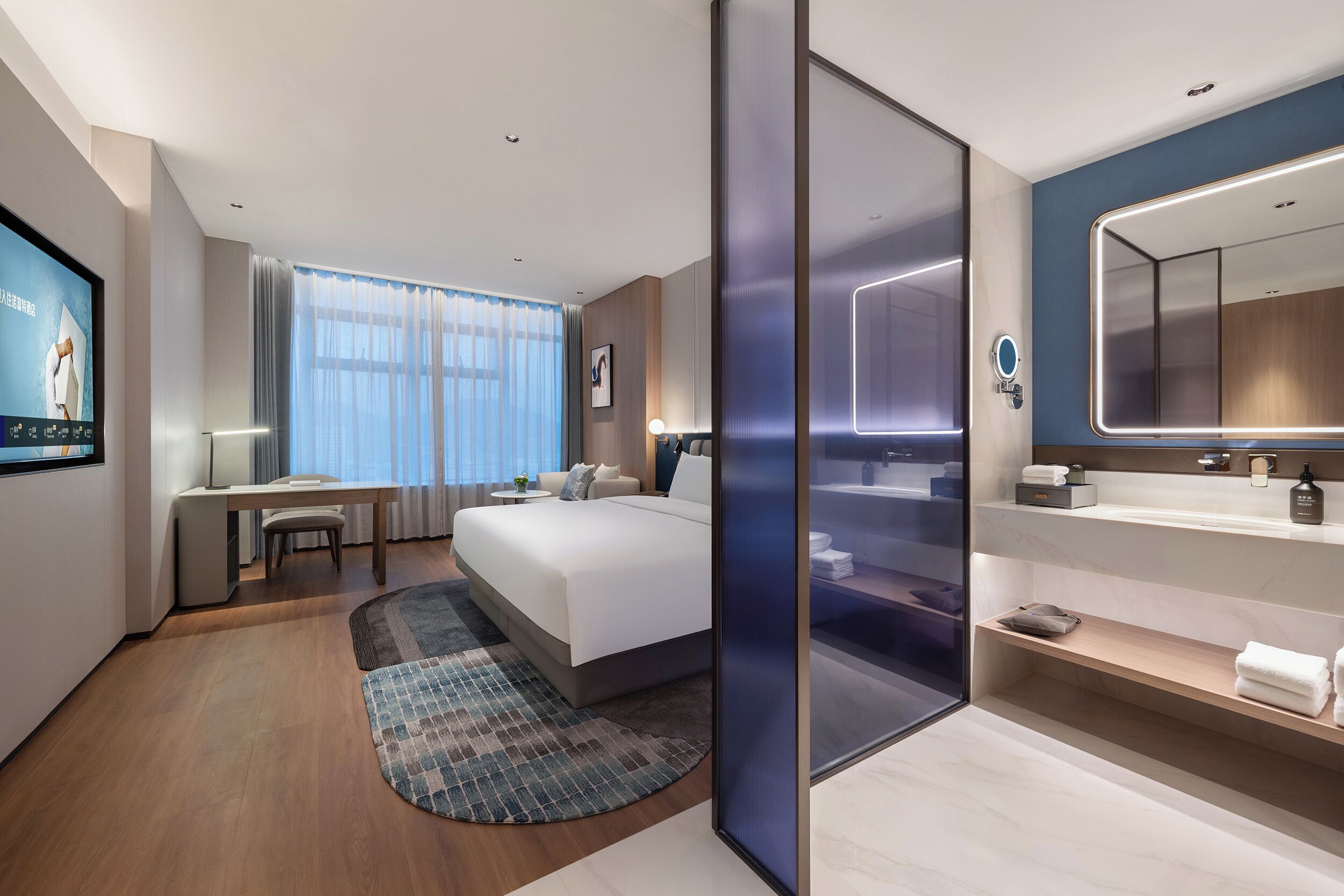 A modern hotel room with a large bed, a desk, a TV, a bathroom with a mirror and shelves, and a view of the city.
