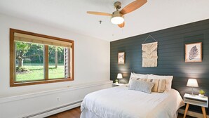 Ein Schlafzimmer mit einem großen Bett, einem Deckenventilator, einem Fenster mit Blick ins Grüne und einer dunklen Wand mit gerahmten Kunstwerken.