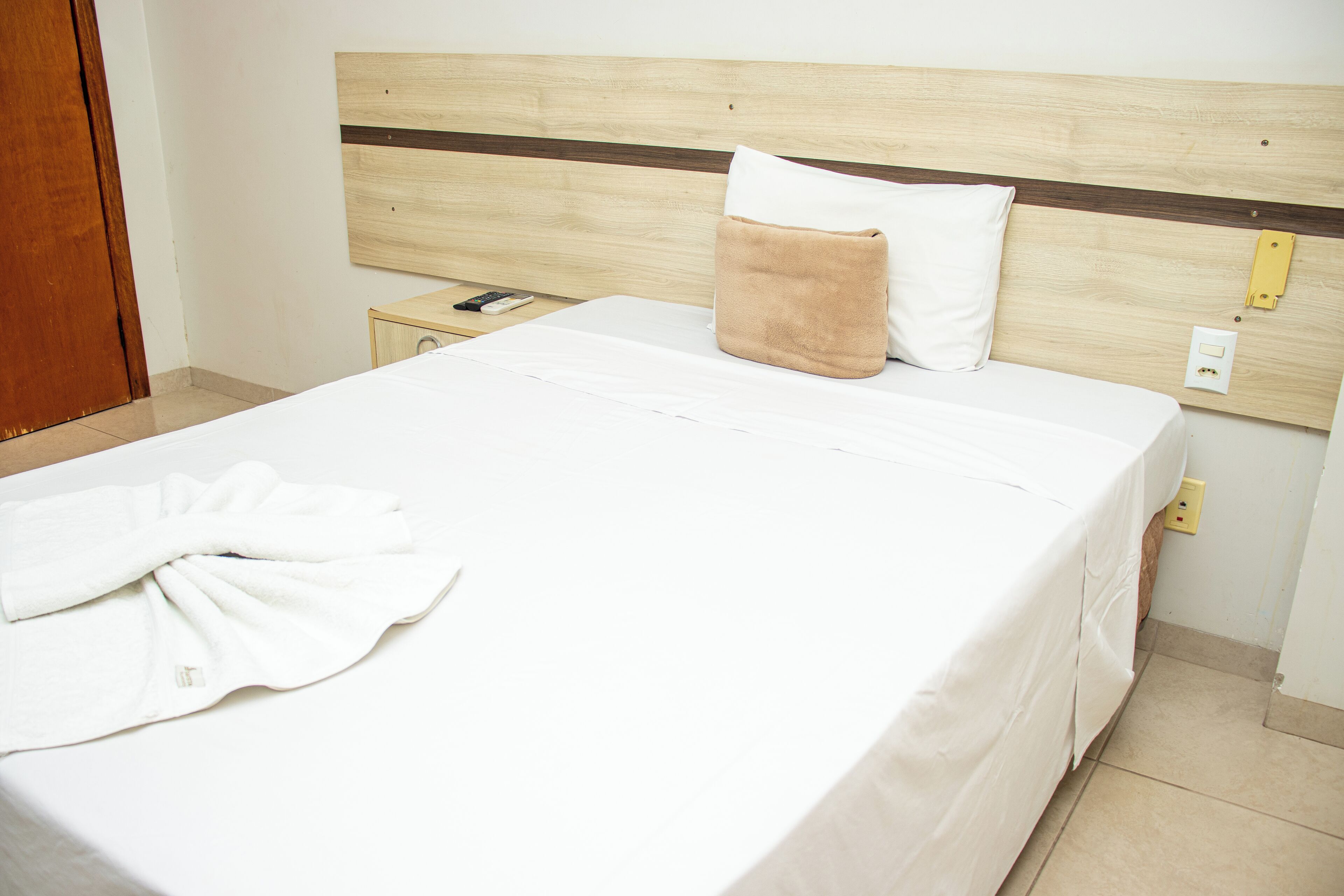 A neatly made bed with a white duvet, a brown pillow, and a white pillow. There is a white bathrobe on the bed and a remote control on the nightstand beside the bed.