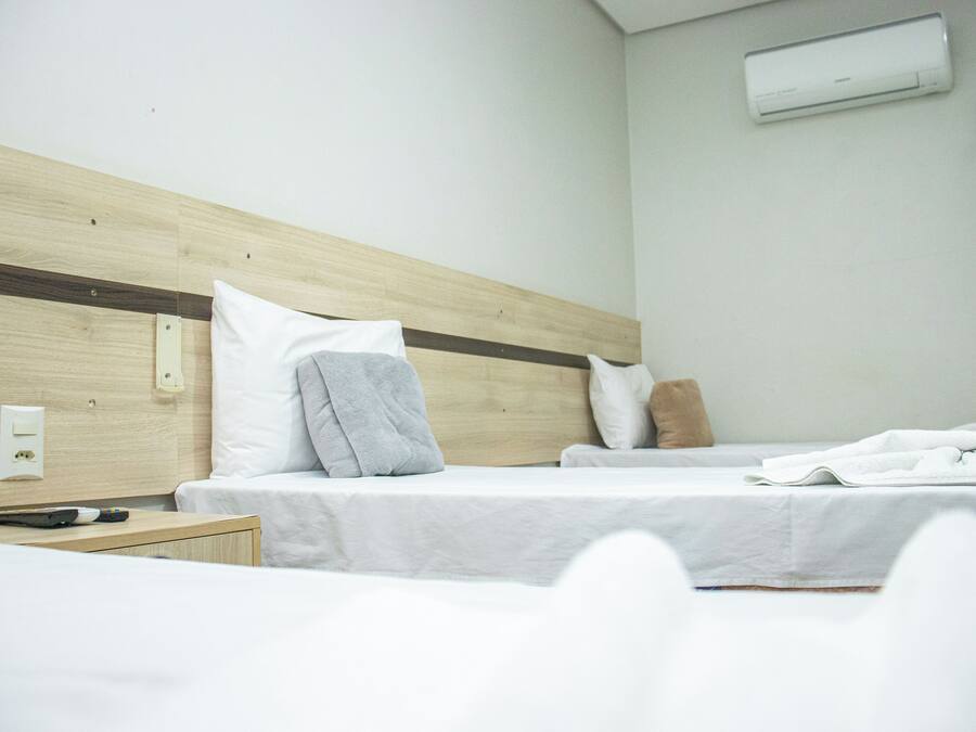 Quarto de hotel com duas camas, cabeceira de madeira, roupa de cama branca, um controle remoto e um aparelho de ar condicionado.