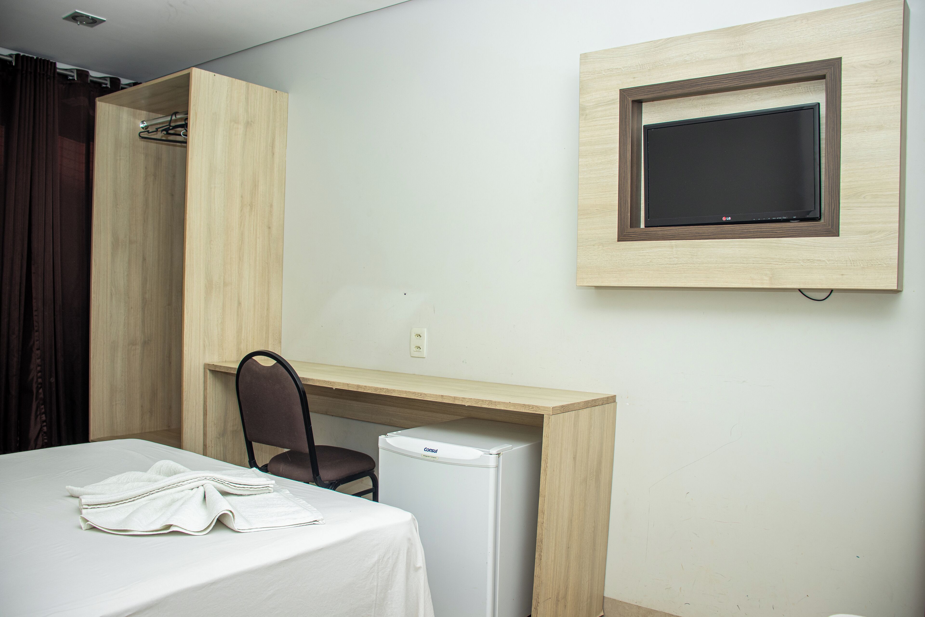 Ett hotellrum med en säng, ett skrivbord, en stol, ett litet kylskåp och en tv som är monterad på väggen.