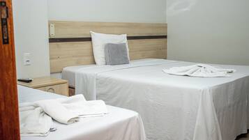 A hotel room with two single beds, each with a white bedspread and a pillow. There is a wooden headboard, a nightstand with a remote control, and a pair of towels on the floor.