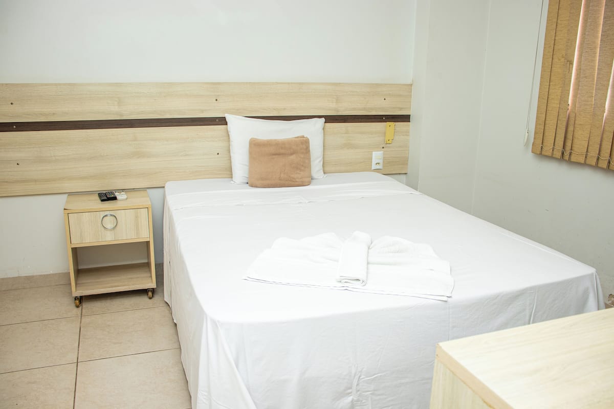 A neatly made bed with white linens and a brown pillow, a wooden nightstand with a remote control, and a wall-mounted light switch.