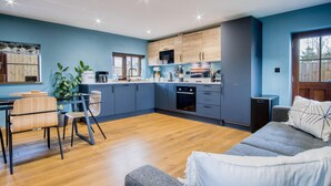 A modern kitchen with blue cabinets, a dining area with a wooden table and chairs, and a grey sofa. - The Old Milking Parlour - 1 Bed Cottage - Amroth (Narberth)