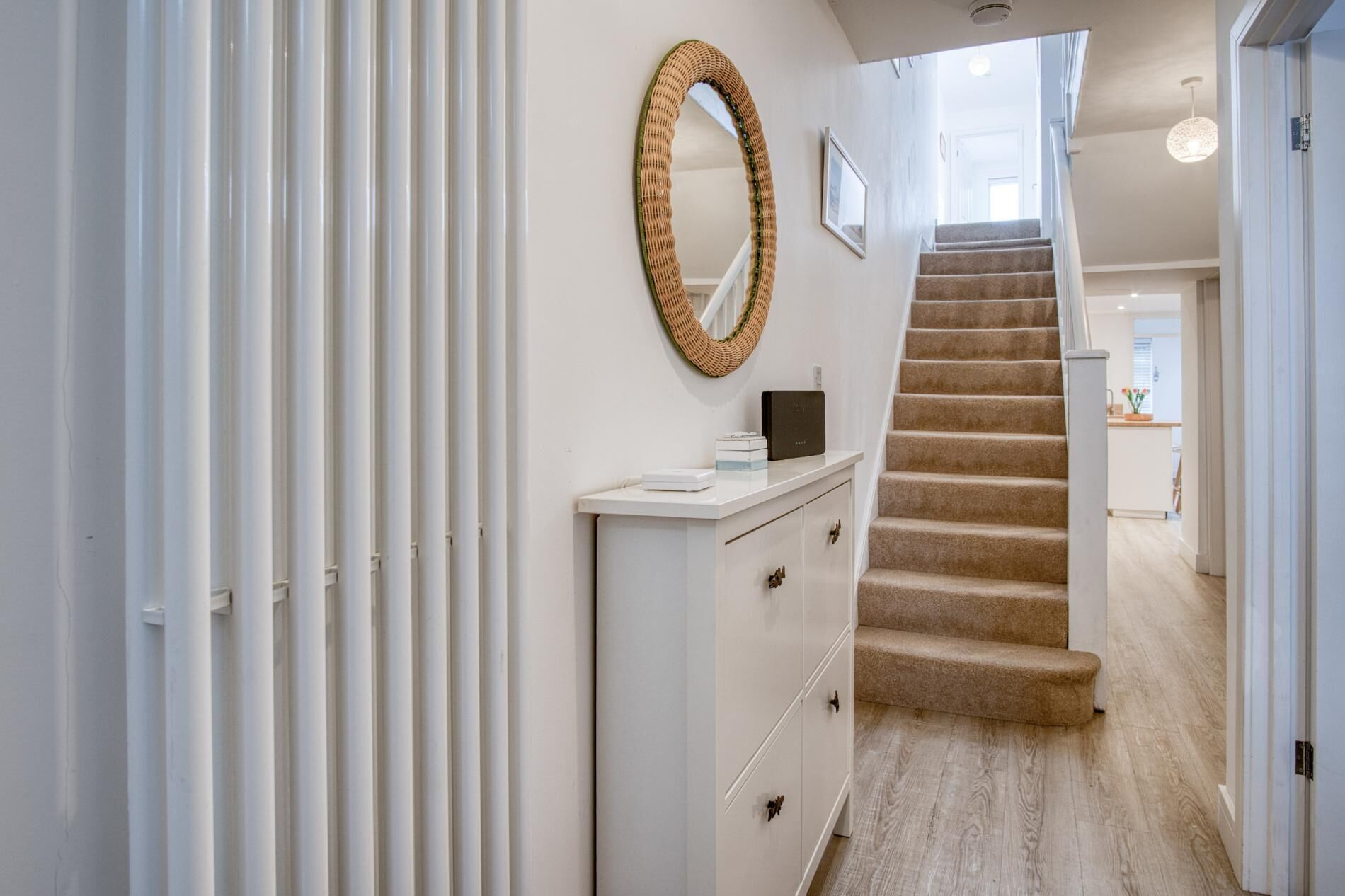 A hallway with a white cabinet, a woven mirror, and a carpeted staircase leading to a kitchen area.