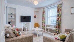 Una sala de estar con dos sofás, un televisor, una mesita con una vela, una ventana con persianas y una cortina colorida.