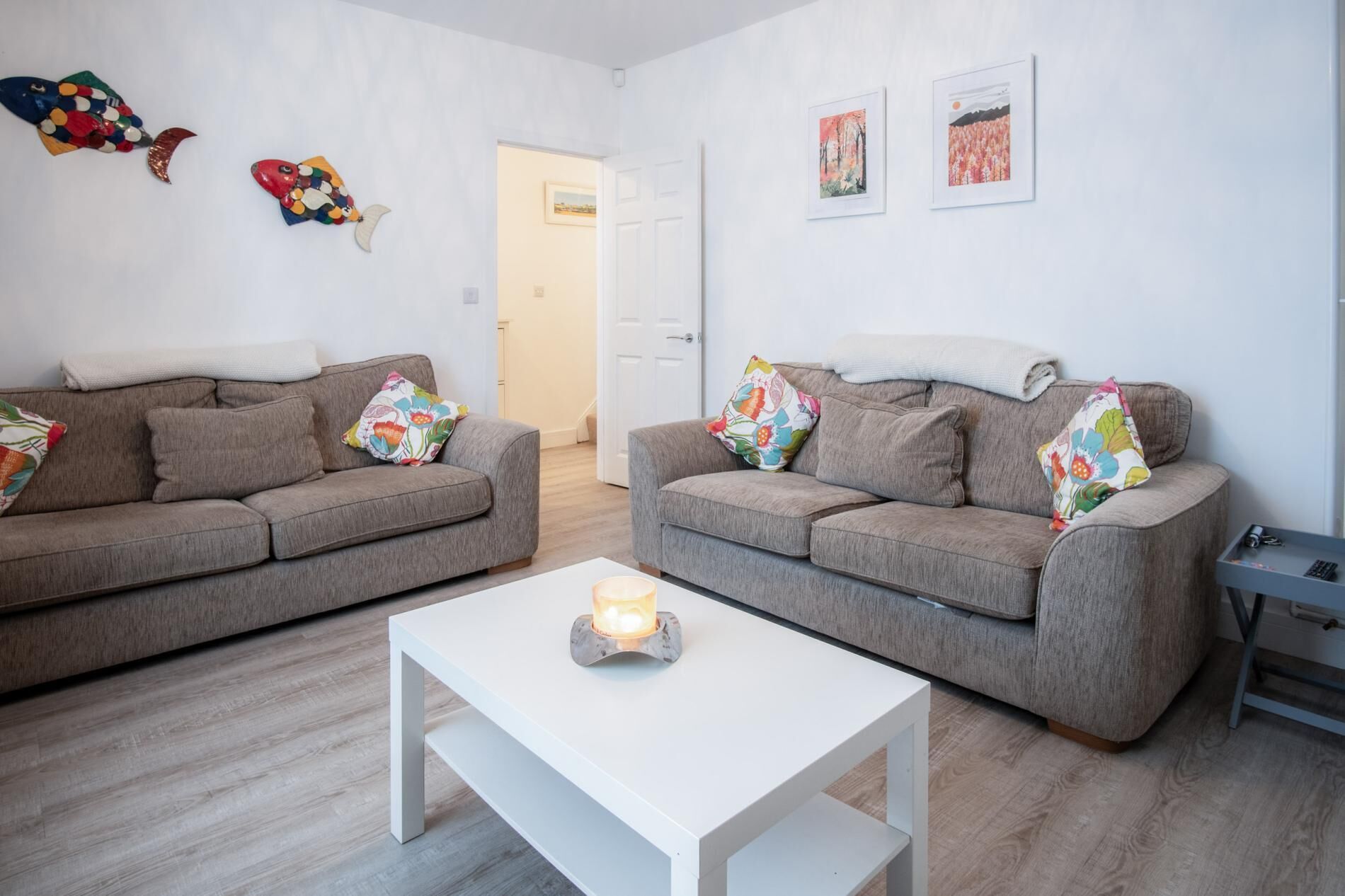 A modern living room with two grey sofas, colorful floral cushions, a white coffee table with a candle, and two framed artworks on the wall.
