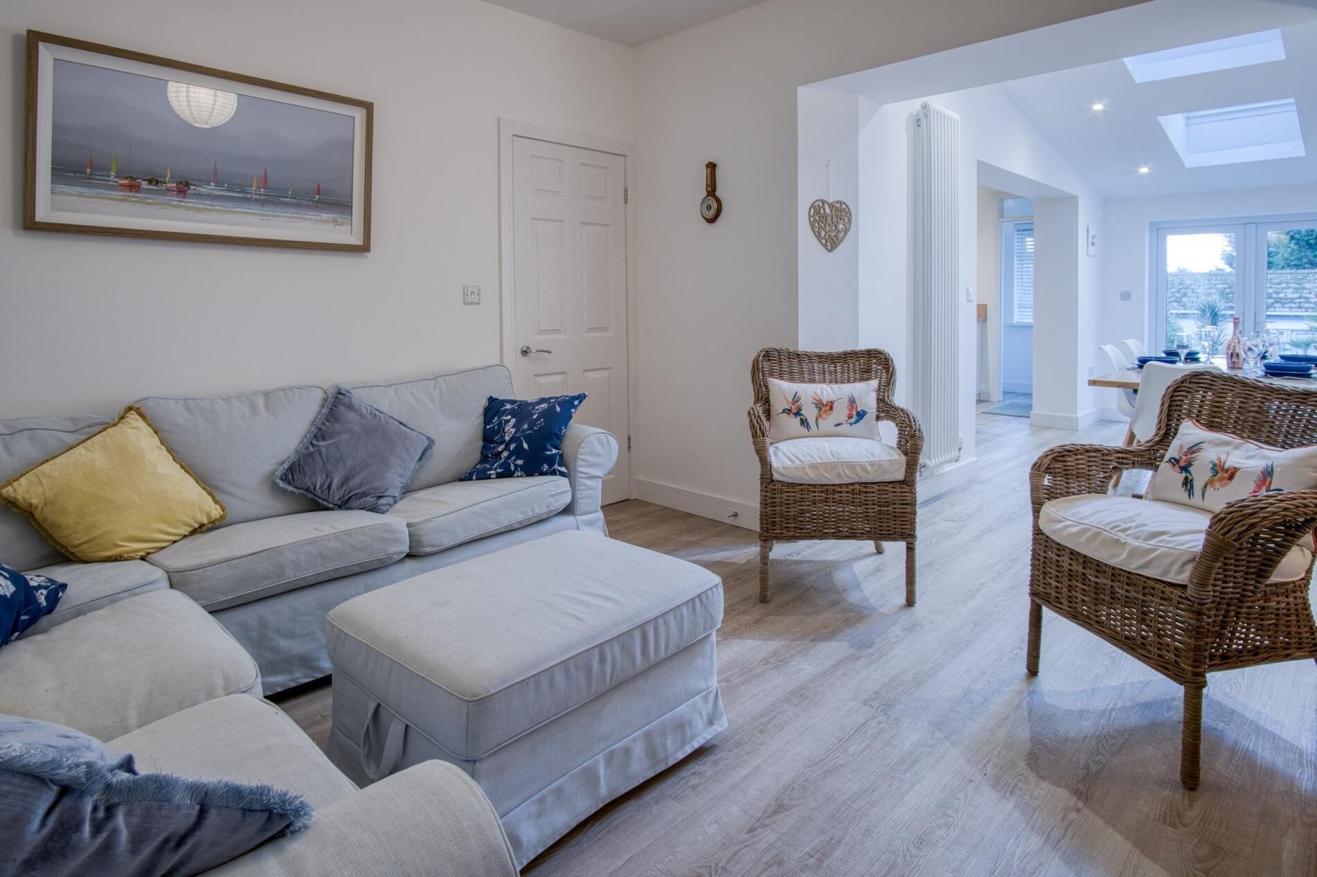 Een woonkamer met een grijze bank, twee rieten stoelen en een witte poef.