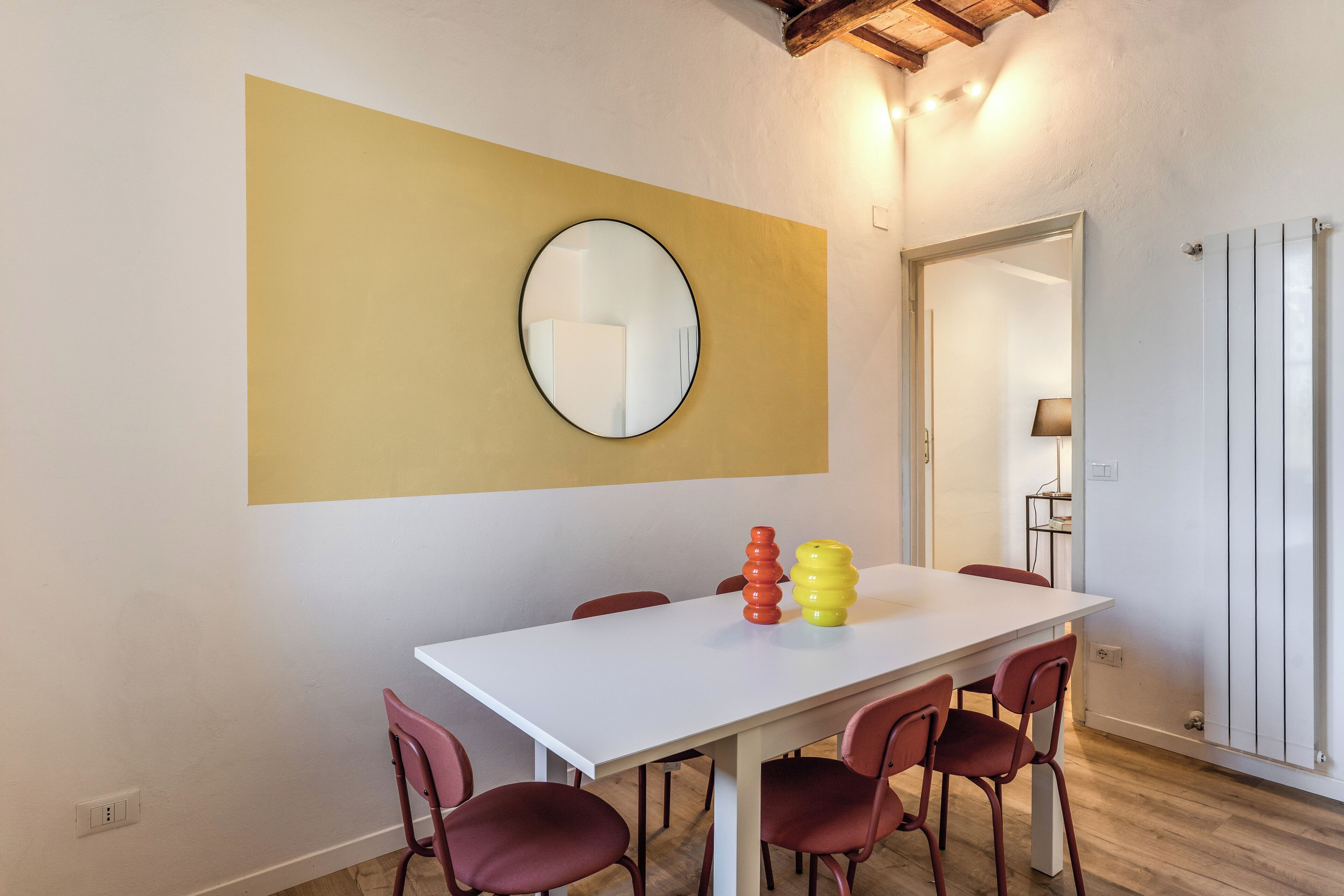 Een eetgedeelte met een witte tafel, rode stoelen, een ronde spiegel en een geel accentmuur.